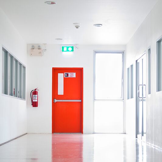 Blick den Gang hinunter auf eine Brandschutztür. Links neben der Tür ein Feuerlöscher.